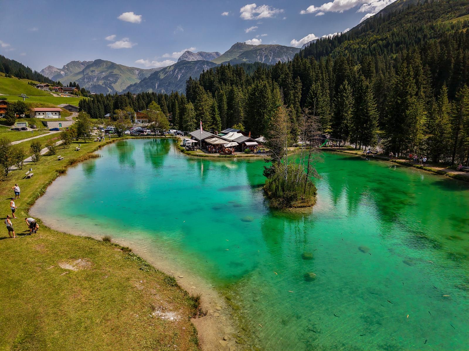Luftaufnahme des türkisgrünen Fischteich Lech mit Restaurant, Insel und Arlberg-Bergpanorama