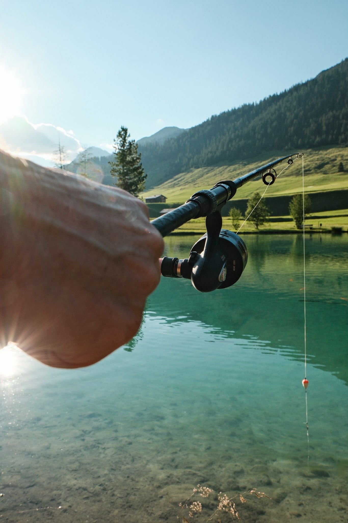Hand mit Angelrute über dem klaren Wasser des Fischteichs, dahinter die Bergwiesen von Zug