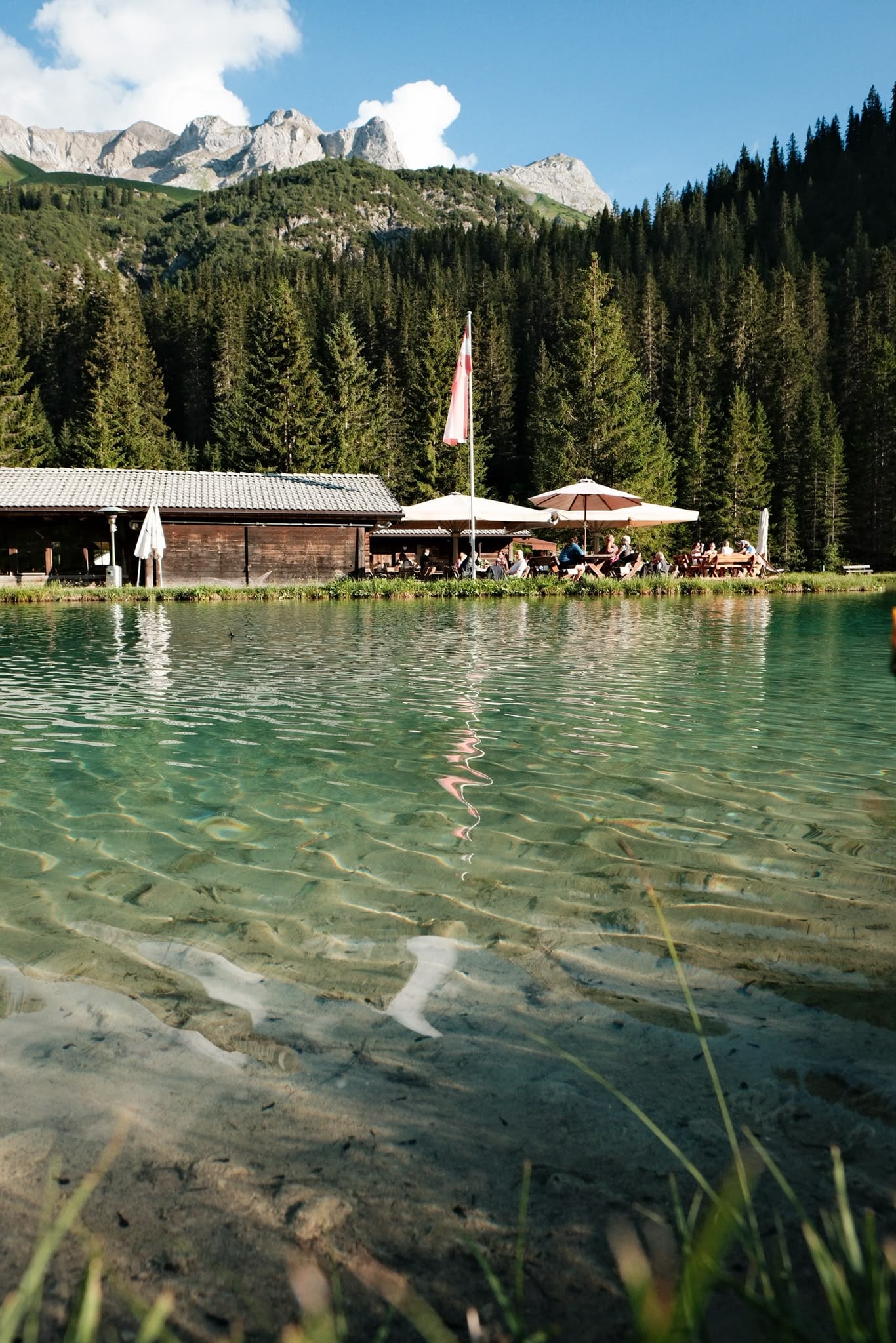 Blick über das klare Wasser des Fischteichs auf das Haus und die Bergflanke dahinter